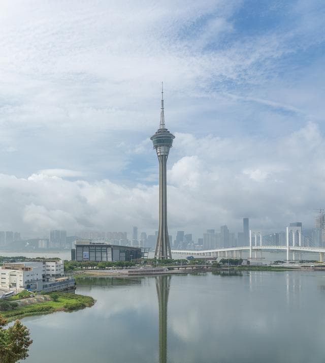 宽阔视角展现澳门塔，作为视觉焦点，周边现代建筑群、白色桥梁与平静水面相映成趣，水面倒映塔影，蓝天缀白云，画面清晰明亮，氛围宁静开阔。